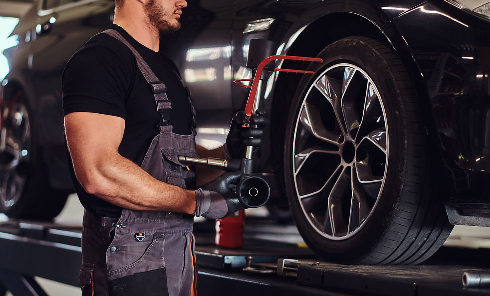 A mechanic in overalls uses a pneumatic tool on a car wheel in a well-lit garage