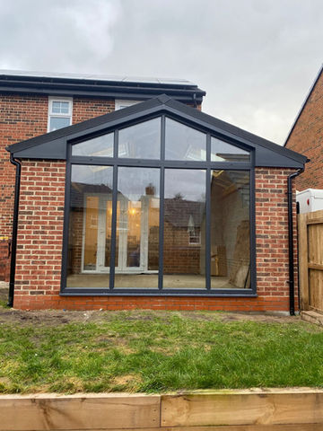 A modern glass conservatory with a gable roof, featuring brick walls and a grassy area
