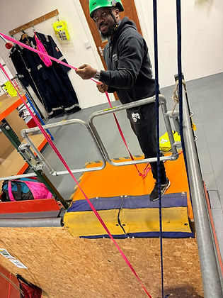 A man wearing a green helmet and black athletic wear holds a pink rope, standing on an orange platform surrounded by safety gear