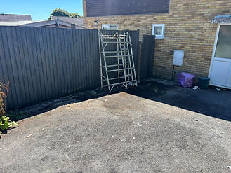 Clean outdoor concrete area beside a brick house with a wooden fence and a stacked wooden frame leaning against it
