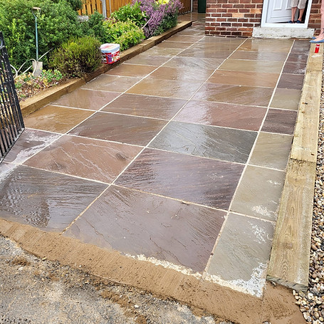 A freshly laid, wet stone patio with rectangular tiles in earthy tones, bordered by a garden