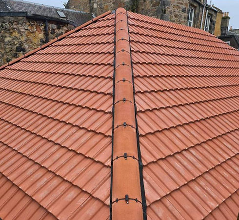 Steeply sloped roof covered with red terracotta tiles, central ridge, stones, and old buildings in the background