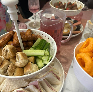 A festive spread with a bowl of snacks including sausage bites, cucumber, pastries, and cheese puffs, along with drinks in the background
