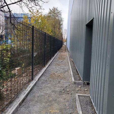 Narrow gravel path between a tall metal fence and a gray building, leading into the distance