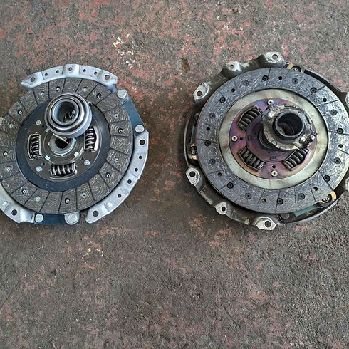 Two car clutch components, one with friction discs and springs, the other with metallic parts, lie on a rough surface