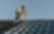 A construction worker sits on a roof, holding a drill, with a wavy metal roofing below