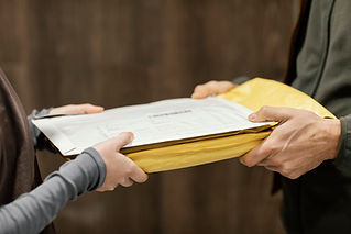 Two hands exchange a yellow padded envelope and a white document against a blurred brown background