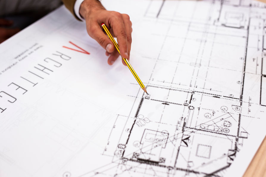 A hand holding a yellow pencil hovers over detailed architectural blueprint