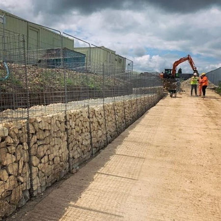 A construction site with a stone-filled wire mesh retaining wall running alongside a dirt path