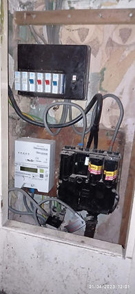 An electrical panel with circuit breakers, wires, and a meter on a worn wall