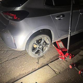 A silver car with a flat rear tire is lifted by a red jack on a dimly lit street