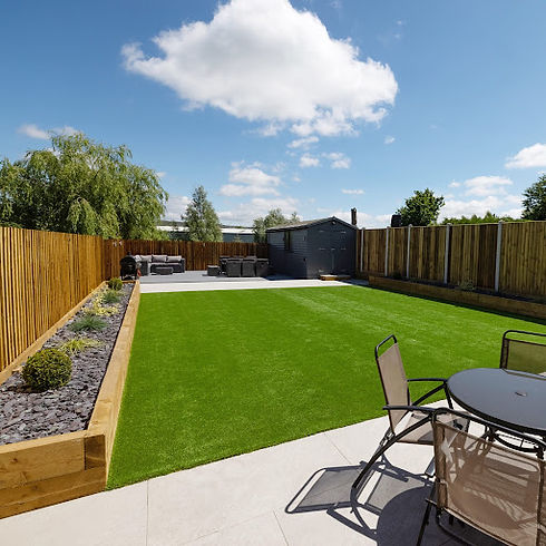 A spacious back garden with green artificial lawn, wooden fences, and a patio area featuring a round table and chairs