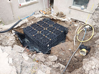 A construction site with a large hole containing a black drainage crate for soakaway installation