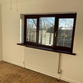 An empty room featuring a large, brown-framed window with two panes, a white radiator below, and wooden flooring