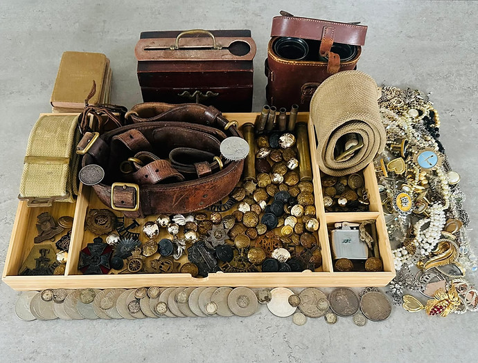 A wooden sorting tray containing antique estate items including silver pocket coins, military uniform buttons, leather service belts, historic insignia badges, and a dense accumulation of vintage costume jewellery. 
