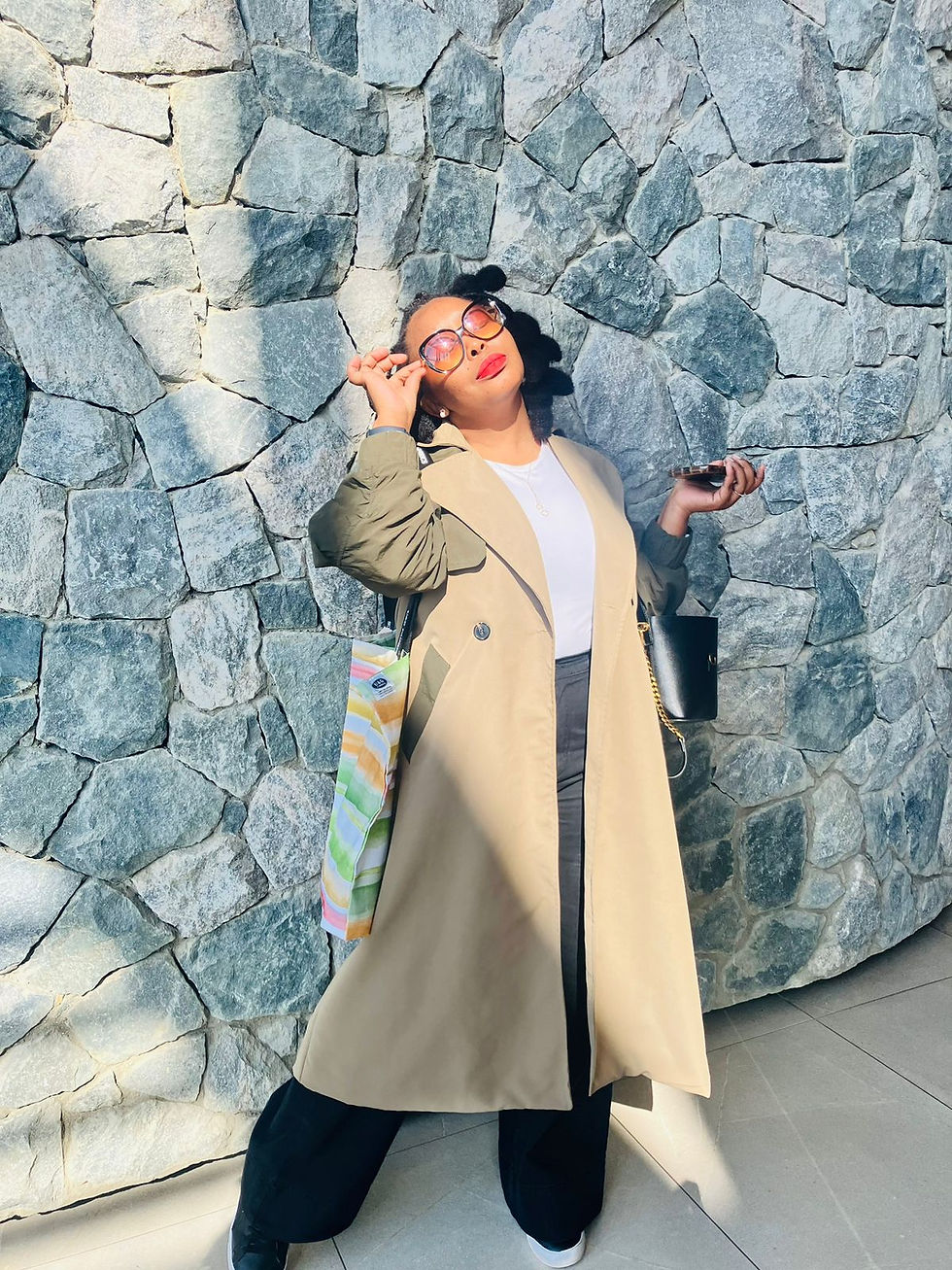Woman in sunglasses poses confidently against a stone wall, wearing a beige coat. She holds a phone and colorful bag, exuding a chic vibe.