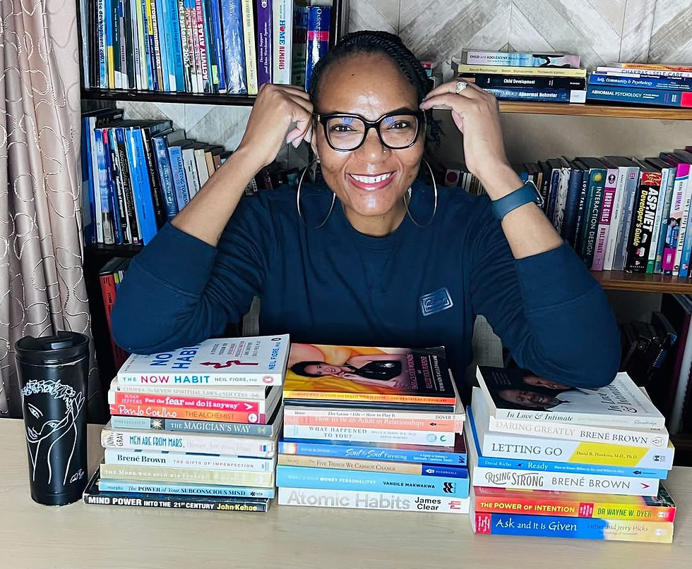 A lady wearing spectacles. She's smiling and surrounded by books. Some books are on a bookshelf whilst her elbows are resting on others.