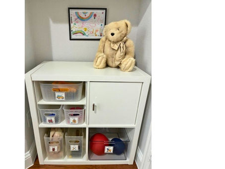 A stuffed teddy bear sitting on top of a Kallax unit that showcases different labeled storage bins that would serve a child well as toy storage.