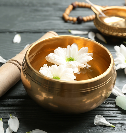 A brass singing bowl filled with water and white flower petals, surrounded by healing crystals and a bamboo stick, creating a peaceful, meditative setting.