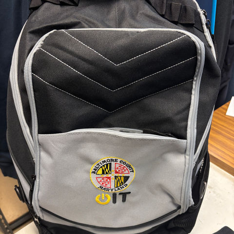 A black and gray travel or day backpack embroidered with a circular custom logo using the colors of the Maryland flag.