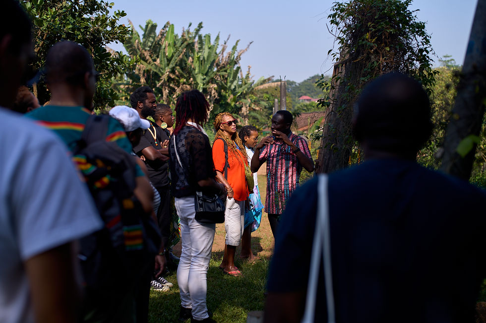 A group of black individuals gathered outdoors in a lush, green environment, listening attentively to a man speaking. The speaker is wearing a colorful patterned shirt, and the group includes individuals in casual clothing, some holding bags or wearing backpacks. Tall trees and vegetation surround the area, suggesting a natural or rural setting. The mood appears educational and engaging, with the group focused on the speaker.