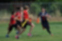 A St. Victor School flag football player in a black Chargers jersey running with the ball while being pursued by players in red jerseys during a game on a grassy field.