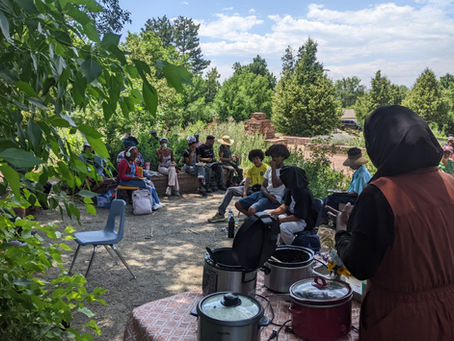 a group gathers in a gravel area, surrounded by greenery. Most of the group have dark hair and skin and are eating and listening to a person speaking in the foreground. The speaker's back is to us and they gesture with a dark-skinned hand. They wear a black hijab, a long sleeve black shirt and a dark-copper vest. There are large pots with lids on a table in front of the speaker. The sunlight dapples the ground and the people in the foreground, while very bright in the area beyond where you can see large trees and a red-stone chimney at the edge of a patio in the same material..