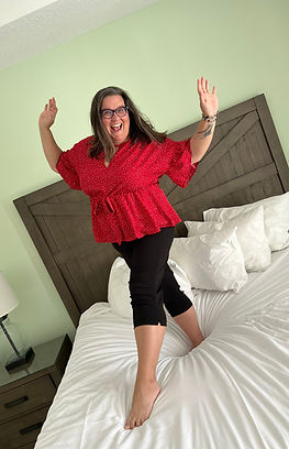 Travel advisor Denise Lang is wearing a red shirt and black pants, laughing while she stands on top of a bed.