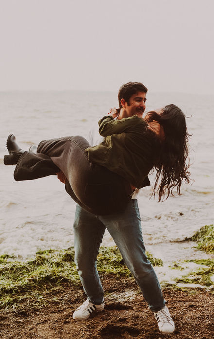 Man holding woman near water; engagement photography capturing loving moment.