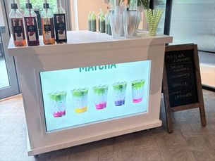 Matcha drink stand with syrup bottles, stacked cups, straws, and a chalkboard listing flavors like salted caramel and strawberry.