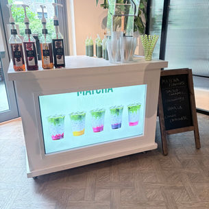 Matcha drink stand with syrup bottles, stacked cups, straws, and a chalkboard listing flavors like salted caramel and strawberry.