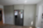 Gray kitchen cabinets and a fridge with magnets and notes. Wooden floor, white walls. A person is peeking into the room on the left.