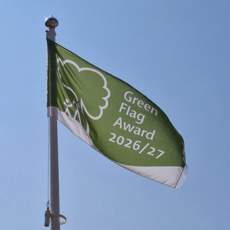 Green flag with 'Green Flag Award 2026/27 in white lettering, and a cartoon image of the tree and adult and child to the left of the wording. Blue sky background.