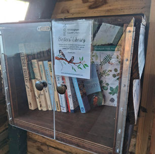 A wooden cupboard on the wall of the bird hide. It has transparent perspex doors with two round brass-effect knobs to open them, Inside there are nine books and a nature identification chart lined up on the shelf.