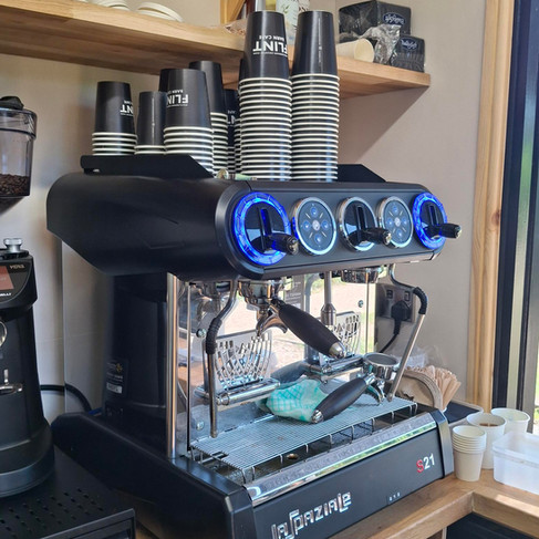 A small, shiny silver coffee machine with Flint Barn Café black take-away cups on top.