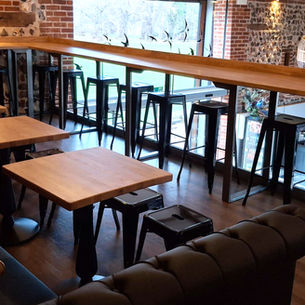 Oak tabletops and an oak bar in the background. In the foreground is the arm of a black bench sofa seating area You can see grass through a window in the background and a large willow woven lampshade above the cafe on the lower level..