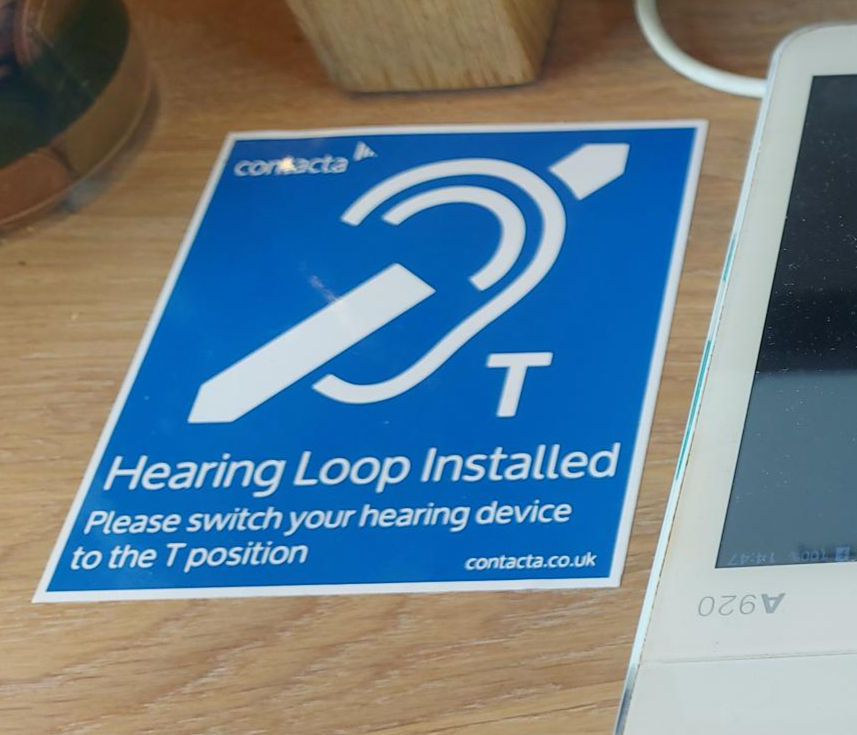 Hearing loop blue sign on the wooden cafe counter. There is an ear icon and letter T in white lettering. The instructions say Hearing Loop Installed - Please switch your hearing device to the T position.
