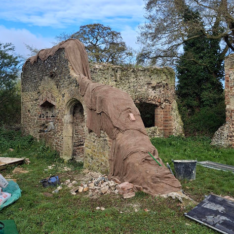 A sloped flint and brick wall with hessian covers to help the lime mortar to set effectively. There are loose flints at the bottom of the wall ready for use.