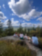 6 children sitting on a boulder overlooking a scenic vista of green trees, mountains, and a vibrant blue sky with puffy white clouds.