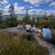 A row of people with their backs to the camera look out over a mountainous forest vista.