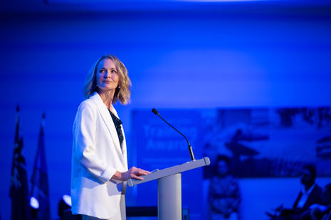 Female speaker in white suit on stage at podium in blue light