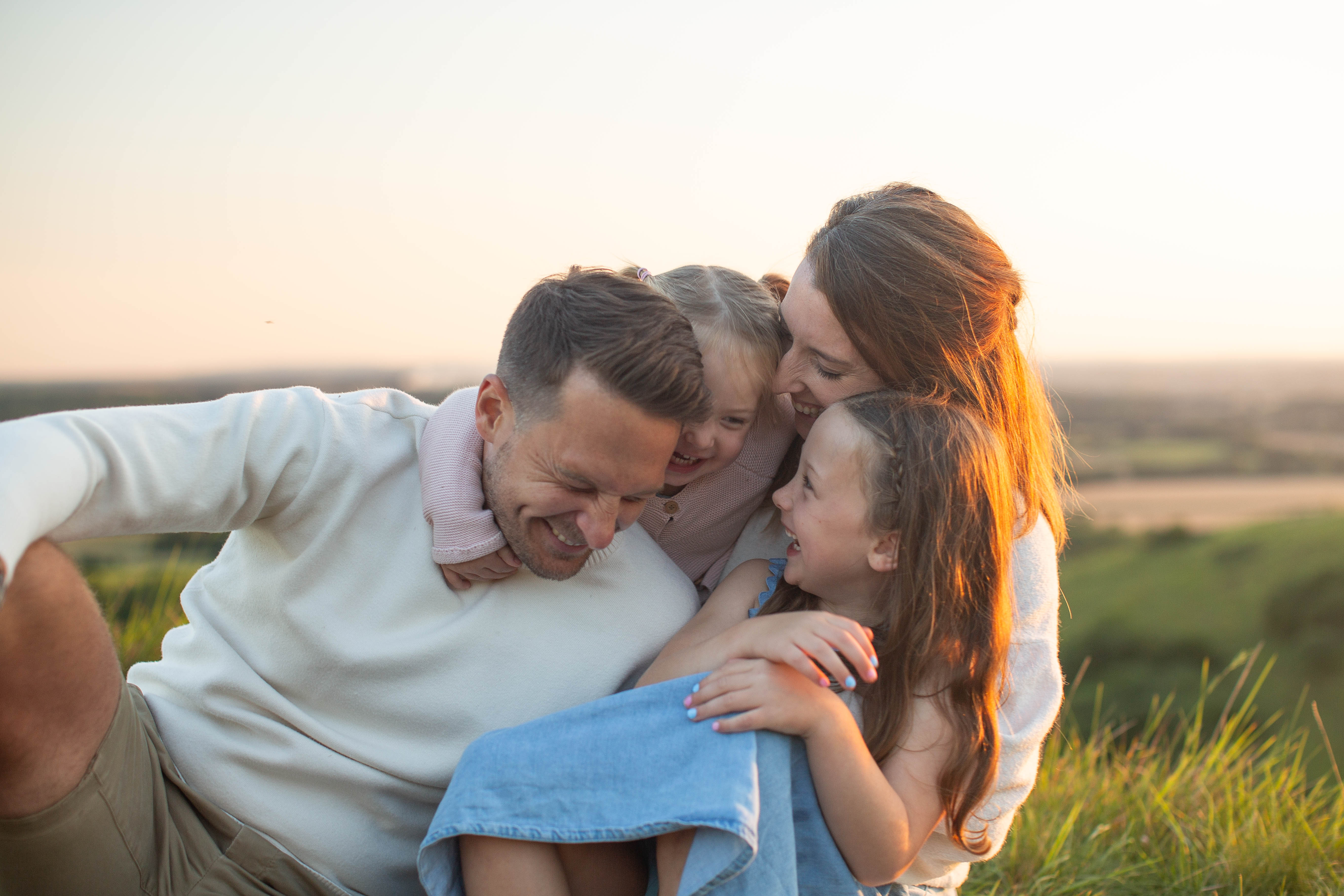 Smiling family of four cuddles on a grassy hill at sunset, laughing together in warm golden light.