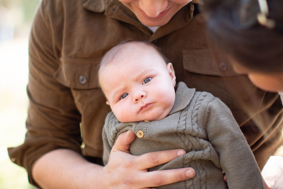 Baby in a brown sweater held by an adult, glancing curiously at another person. Warm, intimate moment outdoors with soft natural light.