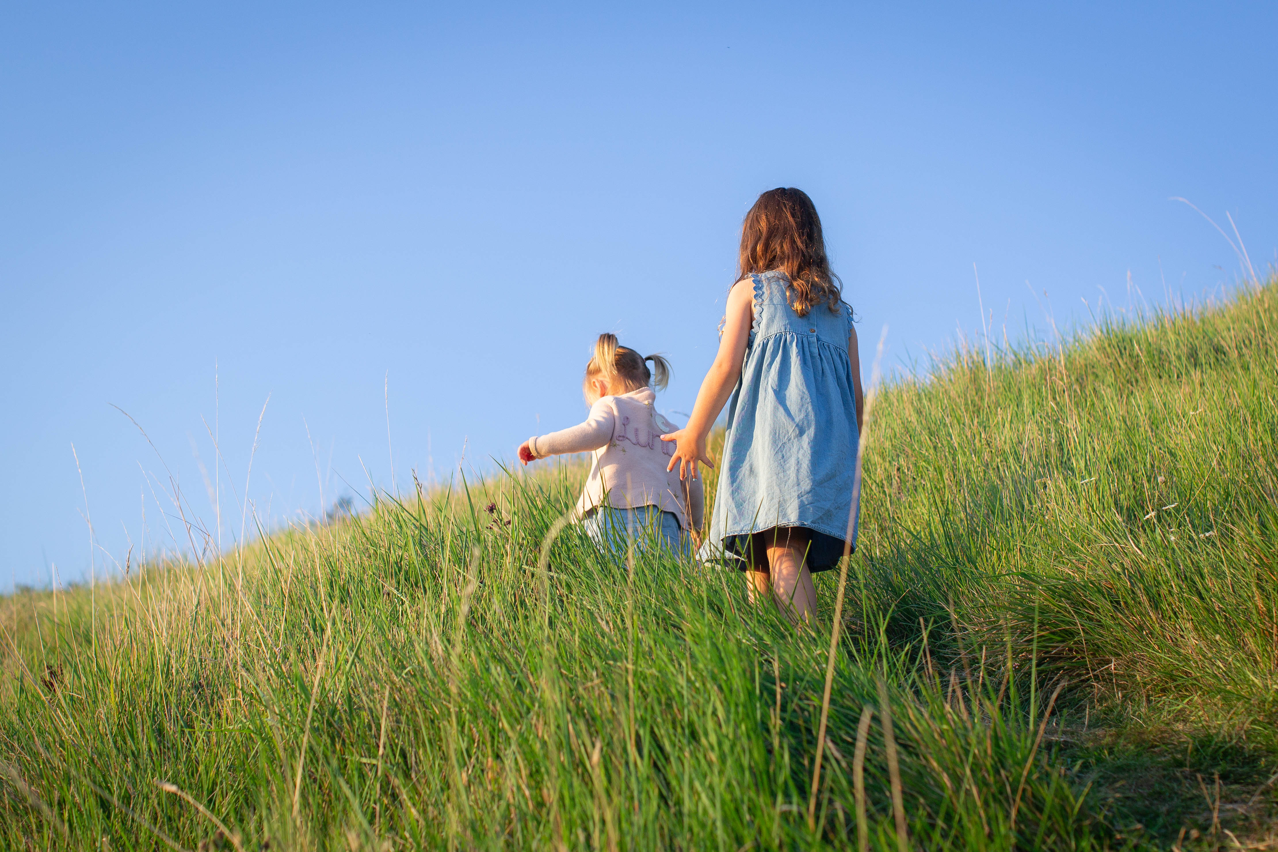 Two young girls walk up a grassy hill under a clear blue sky, one in a denim dress, in a peaceful summer scene
