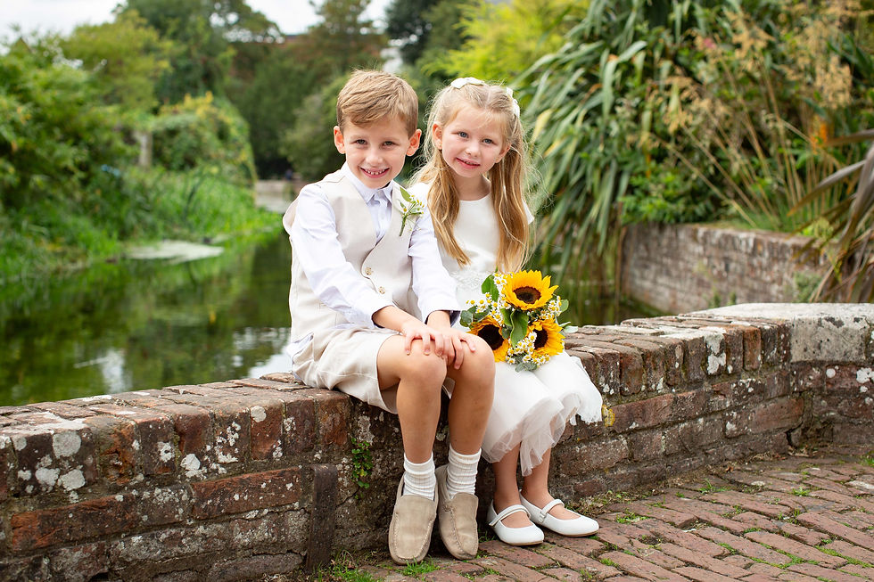 Smiling young boy and girl in white formal clothes sit on a brick wall by a canal, holding a sunflower bouquet in a lush garden.
