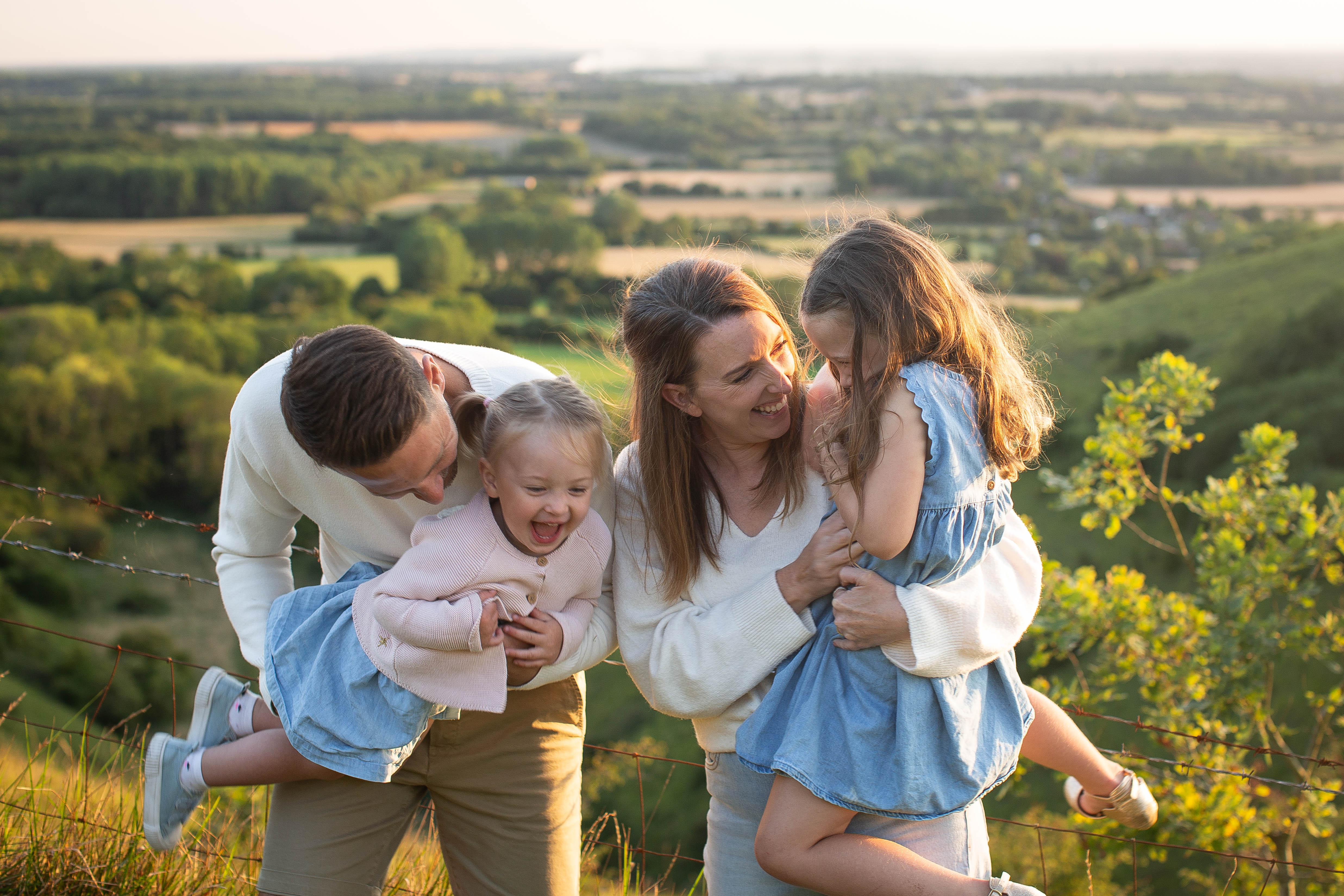 Smiling parents holding two young daughters on a hillside, laughing together above a sunlit green valley at golden hour