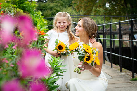 Bride and girl holding sunflowers, smiling outdoors.