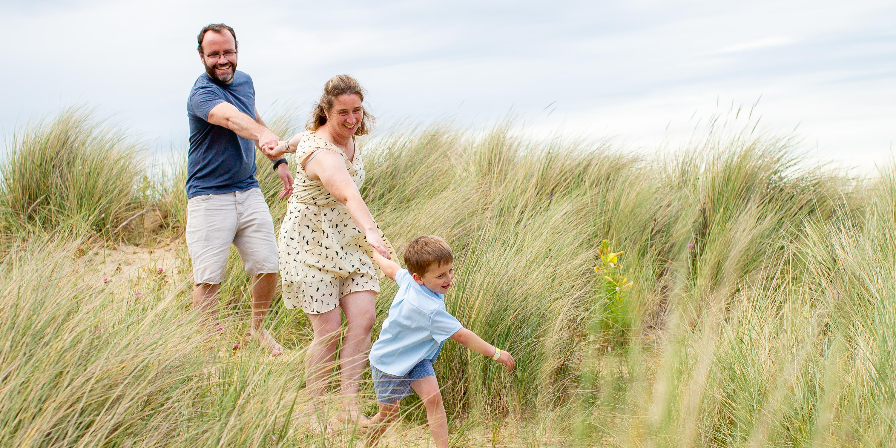 Smiling family of three holding hands and walking through tall beach grass dunes on a windy day.