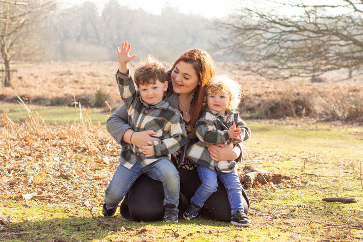 Smiling woman kneels in a sunny field, hugging two young children in plaid shirts; one waves, autumn trees behind.