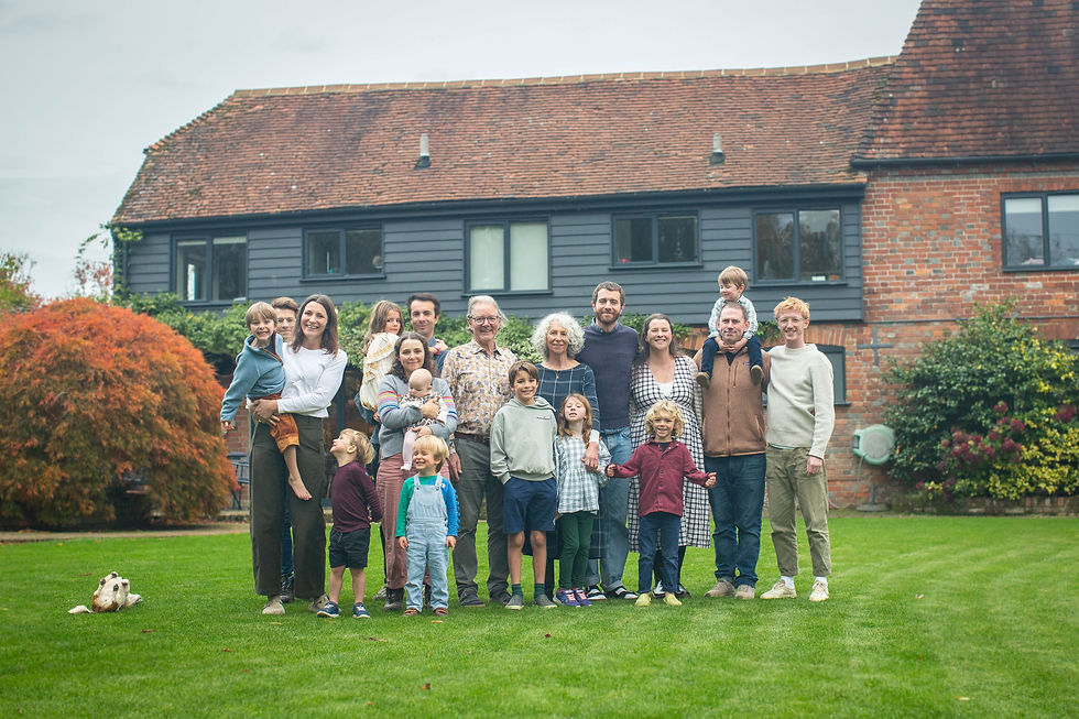 A large family poses happily on a green lawn in front of a red-brick house, with a dog lying nearby and autumn foliage in the background.